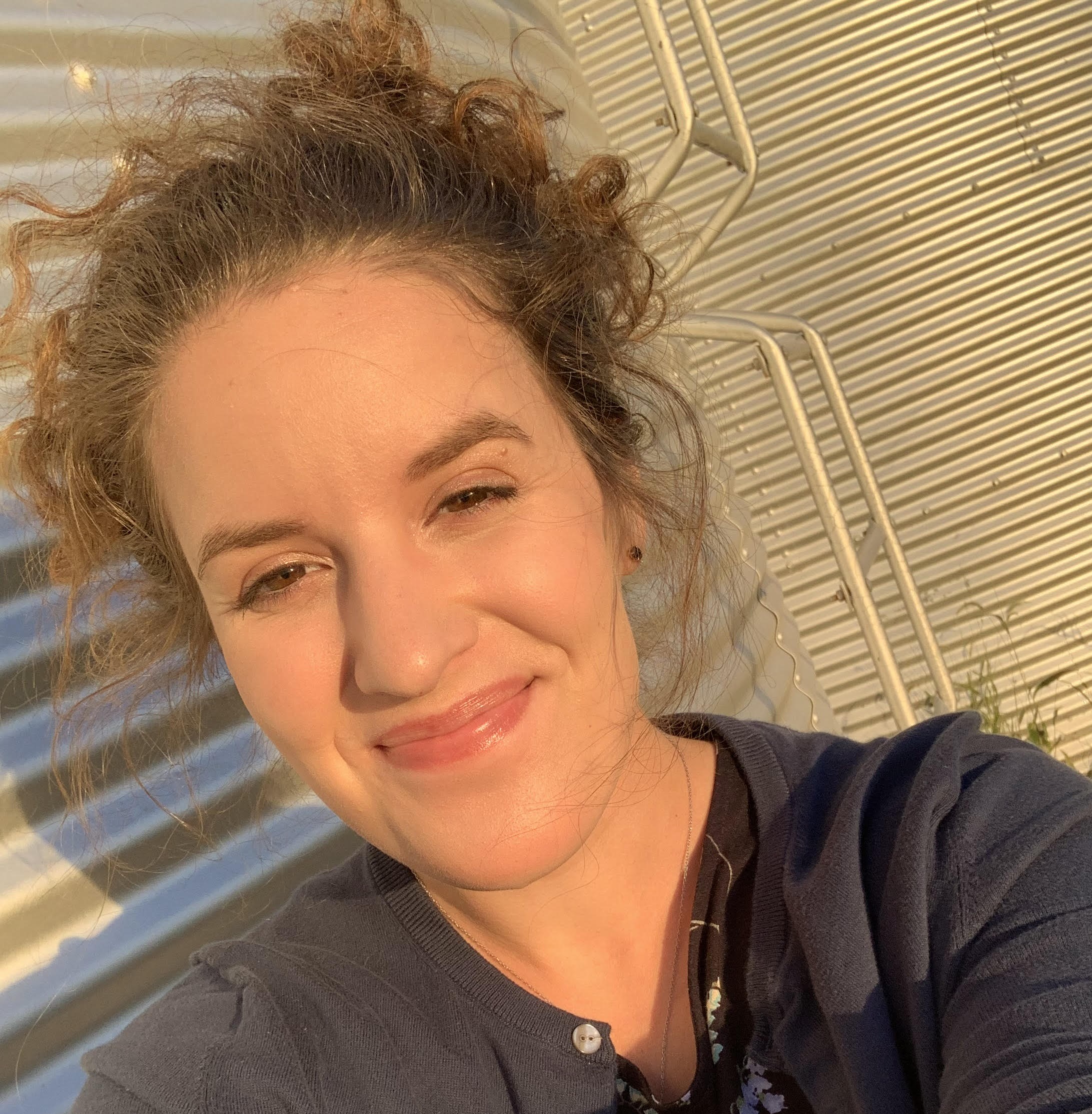 Selfie of me smiling at the camera. I have curly brown hair and brown eyes and I am wearing a grey t-shirt with a grey cardigan.