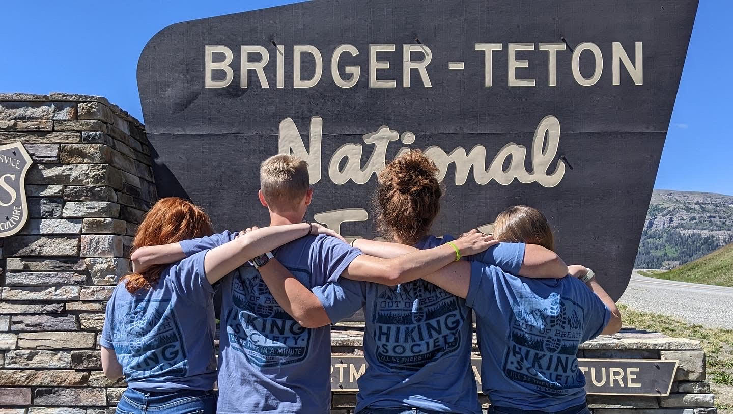 Me and three of my friends in front of the Bridger-Teton National Park sign. We are wearing matching blue tshirts that say Out of Breath Hiking Society.